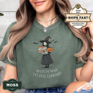 May include: A green t-shirt with a cartoon witch carrying a stack of books and a pumpkin. The text "Witch Way To The Library" is printed on the shirt.