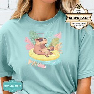 May include: A light blue t-shirt with a graphic of a capybara wearing sunglasses and a floatie in a tropical setting. The text "Ships Fast" is on a green and white sticker.