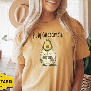 A humorous t-shirt design featuring an avocado dressed as a monk with a cross belt, perfect for food humor enthusiasts. Ideal for those who love avocado puns, funny Christian gifts, and funny phrase slogans for a unique twist on Holy Guacamole.