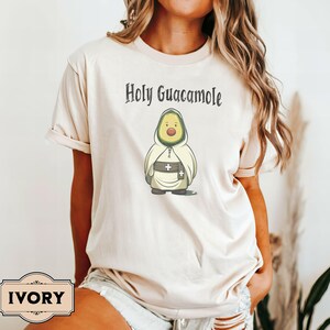 A humorous t-shirt design featuring an avocado dressed as a monk with a cross belt, perfect for food humor enthusiasts. Ideal for those who love avocado puns, funny Christian gifts, and funny phrase slogans for a unique twist on Holy Guacamole.