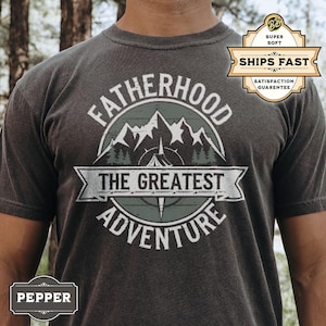 May include: A dark grey t-shirt with a graphic design of a compass and mountains. The text "Fatherhood The Greatest Adventure" is printed on the shirt.