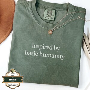 May include: An olive green t-shirt with the text "inspired by basic humanity" in white. A gold necklace with a pendant rests on the shirt. The shirt has a Comfort Colors tag. A brown hat and dried palm leaves are in the background.