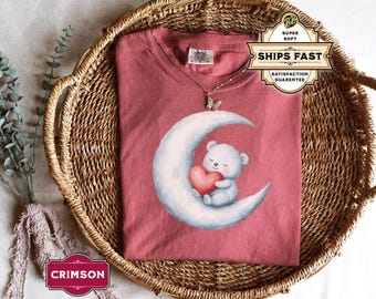 Op de afbeelding: Een koraalrood T-shirt met een witte maan en een witte beer die een rood hart vasthoudt. De beer zit op de maan. Het T-shirt ligt in een gevlochten mand.