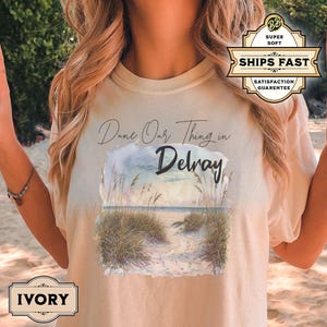 May include: A beige t-shirt with a watercolor illustration of a beach scene and the text "Dune Our Thing in Delray".