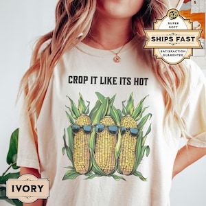 May include: Ivory t-shirt with a graphic of three corn cobs wearing sunglasses, set against green leaves. The text "CROP IT LIKE ITS HOT" is above the corn. The shirt has a loose fit.