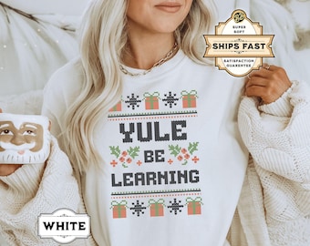 May include: White Christmas sweater with a red, green, and white design. The sweater has the text "Yule Be Learning" on it.