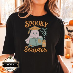 May include: Black t-shirt with a Halloween graphic. The design features a cartoon ghost wearing a mortarboard, reading a book, and the words "Spooky Scripter". A small bottle and bats are also included in the design.