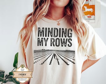 Op de afbeelding: Crèmekleurig T-shirt met de tekst "MINDING MY ROWS" in vette, zwarte letters. Daaronder een zwart-wit illustratie van een persoon die door een veld loopt. Het shirt heeft een relaxte pasvorm.