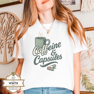 May include: White t-shirt featuring the text "Caffeine & Capsules" with a coffee mug and capsules graphic. The design is in a stylized font. The shirt is being worn by a person.