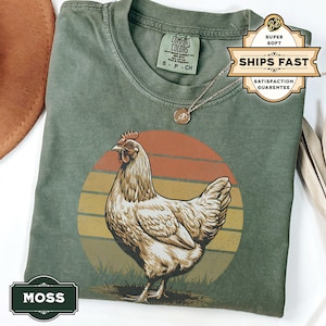 May include: A green t-shirt with a vintage-style illustration of a chicken in front of a sunset. The text "MOSS" is printed on the bottom left corner of the shirt.