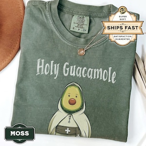 A humorous t-shirt design featuring an avocado dressed as a monk with a cross belt, perfect for food humor enthusiasts. Ideal for those who love avocado puns, funny Christian gifts, and funny phrase slogans for a unique twist on Holy Guacamole.