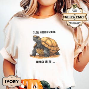 May include: A white t-shirt with a graphic of a turtle carrying a jack-o'-lantern. The text "Slow Motion Spook" and "Almost There..." is printed on the shirt.