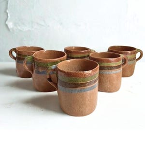 May include: Six brown ceramic mugs with handles. Each mug features horizontal stripes in shades of green, orange, and blue. The mugs are arranged on a white surface, showcasing their rustic design and earthy tones.