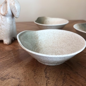Könnte beinhalten: Drei gesprenkelte Keramikschalen mit einer einzigartigen, asymmetrischen Form. Die Schalen haben ein zweifarbiges Farbschema, das von hellem Beige zu gedämpftem Grün übergeht. Eine Keramik-Schaf-Figur befindet sich im Hintergrund.