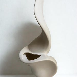 Puede incluir: Escultura de cerámica beige, abstracta, con una forma fluida y orgánica. La escultura tiene un acabado liso y mate y presenta una forma curva y alargada con una abertura central. Diseño moderno y artístico, adecuado para la decoración del hogar.