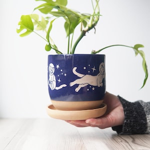 May include: A cylindrical ceramic plant pot with a deep blue glazed upper section featuring white illustrations of cats wearing astronaut helmets surrounded by stars. The lower portion and the matching circular saucer are unglazed tan clay.