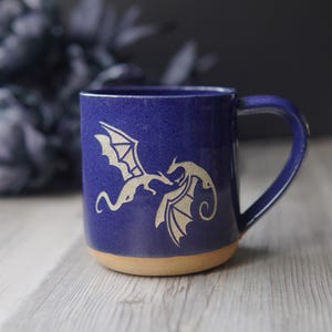 May include: A deep blue ceramic mug featuring a light-colored etched design of two dragons facing each other. The mug has a smooth, glossy finish on the upper portion and an unglazed, natural tan clay base with a sturdy handle on the right side.