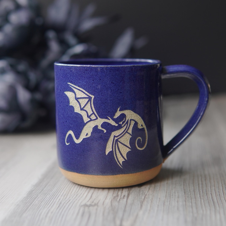 May include: A deep blue ceramic mug featuring a light-colored etched design of two dragons facing each other. The mug has a smooth, glossy finish on the upper portion and an unglazed, natural tan clay base with a sturdy handle on the right side.