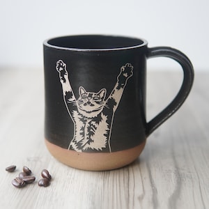 May include: Black ceramic coffee mug with a brown rim. The mug features a white silhouette of a cat with its arms raised in the air.