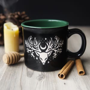 May include: Black ceramic mug with a green rim, featuring a white deer head design with antlers, a crescent moon, and stars. The mug is set on a wooden surface with a lit candle, honey dipper, and cinnamon sticks.