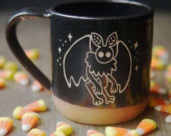 May include: Black ceramic mug with a white design of a creature with wings and large eyes. The mug has a brown rim and is surrounded by candy corn.