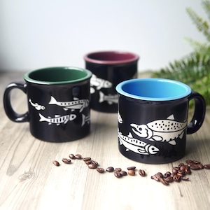 May include: Three black ceramic mugs featuring white fish illustrations. Each mug has a distinct interior colour: one green, one maroon, and one light blue. The mugs are arranged on a wooden surface with scattered roasted coffee beans nearby.