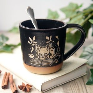 May include: Black ceramic mug with a tan rim and handle. The mug features a white illustration of a cat wearing glasses and reading a book.