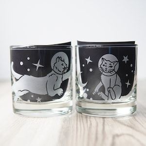 May include: Two clear glass tumblers with black interiors. Each tumbler features a white silhouette of a cat wearing a space helmet and floating in space. The cat on the left is facing right and the cat on the right is facing left. Both cats have a star and a crescent moon behind them.