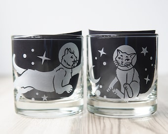 May include: Two clear glass tumblers featuring etched designs of cats wearing space helmets. The left glass shows a cat floating horizontally, while the right glass shows a cat sitting upright. Both designs include stars and circular shapes.