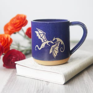 May include: A deep blue ceramic mug featuring a gold-colored graphic of two dragons facing each other. The mug has a smooth, glazed finish on the upper portion and an unglazed, tan-colored base. It includes a curved handle on the right side.