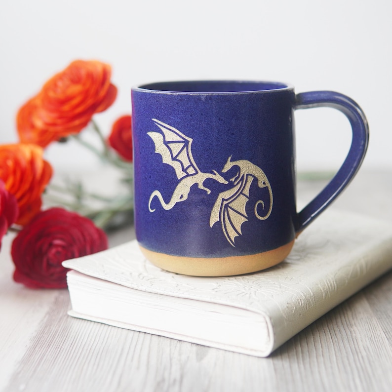 May include: A deep blue ceramic mug featuring a gold-colored graphic of two dragons facing each other. The mug has a smooth, glazed finish on the upper portion and an unglazed, tan-colored base. It includes a curved handle on the right side.