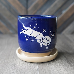 May include: A deep blue ceramic planter with a white graphic of a dog wearing a space helmet, surrounded by stars and a crescent moon. The cylindrical pot features a smooth glazed finish on the upper portion and a natural beige unglazed base.