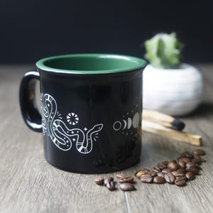 May include: Black ceramic mug with a green interior rim, featuring a white snake and moon phase design. The mug is on a wooden surface with coffee beans, a cactus, and a smudge stick.