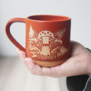 May include: A terracotta mug with a white design of mushrooms and ferns. The mug has a rounded handle and a slightly flared rim.