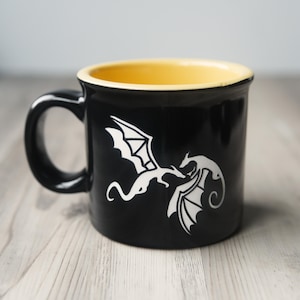 May include: A black ceramic mug with a yellow interior and a handle on the left side. The exterior features a white graphic illustration of two dragons facing each other in flight, with wings spread and tails curved, centered on the front surface.