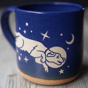 May include: A blue ceramic mug with a white silhouette of a dog wearing a space helmet and flying through space. The mug is decorated with stars and a crescent moon.