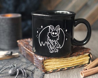 May include: A black ceramic mug featuring a white line art illustration of a moth-like creature with large wings, antennae, and small stars surrounding it. The mug has a rounded handle and a smooth, glossy finish, displaying a clean graphic design.