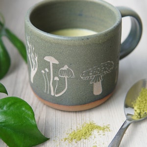 May include: A green ceramic mug with a white mushroom design. The mug is filled with a light green liquid and there is a small pile of green powder on a white surface.