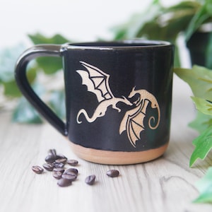 May include: A black ceramic mug featuring a light tan engraved design of two dragons facing each other. The mug has a smooth, glossy black finish on the upper portion and a natural, unglazed tan clay base at the bottom with a curved handle.