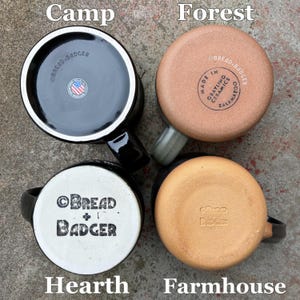 May include: Four ceramic mugs viewed from the bottom. The top left mug has a black base with a circular sticker featuring a US flag. The top right mug has a tan, unglazed base with text. The bottom left has a white base with black text. The bottom right has a tan base with embossed text.