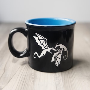 May include: A black ceramic mug featuring a white graphic of two dragons facing each other. The mug has a glossy black exterior and a bright blue interior. The handle is curved and black, matching the exterior finish of the cylindrical vessel.