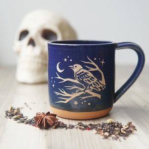 May include: A dark blue ceramic mug with a raven and celestial design. The mug features a raven perched on a branch, a crescent moon, and stars. The mug has a brown base and a curved handle. A skull is blurred in the background.
