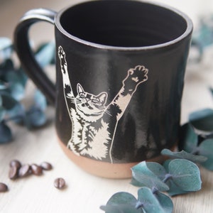 May include: Black ceramic coffee mug with a white silhouette of a cat stretching its arms up. The mug has a brown rim and base.
