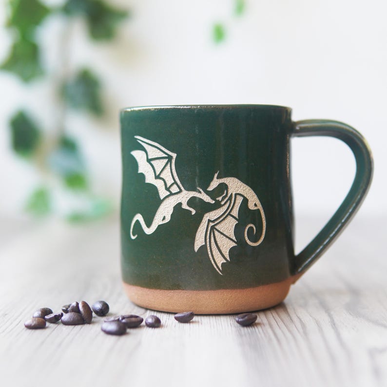 May include: A dark green ceramic mug featuring a light-colored etched design of two dragons facing each other. The mug has a smooth glazed upper section and an unglazed, textured terracotta-colored base, with a single handle on the right side.