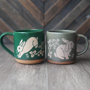 May include: Two ceramic mugs with unglazed terracotta bases. The left mug is vibrant green with a cream-colored leaping rabbit and berry vine design. The right mug is muted sage green with a cream-colored sitting rabbit and berry vine design.