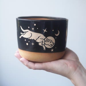 May include: A black ceramic mug with a natural tan base featuring a carved graphic of a dog wearing a space helmet floating among stars and a crescent moon. The design is etched into the dark surface, revealing the light tan clay color underneath.