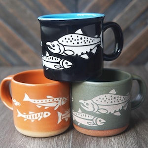 May include: Three ceramic mugs featuring fish illustrations. The top mug is black with a bright blue interior. The bottom left mug is orange, and the bottom right mug is olive green. Each mug displays white or light-coloured fish motifs.