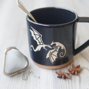 May include: A black ceramic mug featuring an etched design of two dragons facing each other. The mug has a handle, a natural clay-colored base, and contains a silver spoon. A triangular metal tea infuser and two star anise pods are nearby.