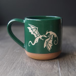 May include: A forest green ceramic mug featuring a light-colored etched design of two flying dragons facing each other. The mug has a smooth glazed upper section and a contrasting unglazed terracotta-colored base with a curved handle on the side.