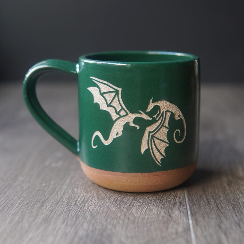 May include: A forest green ceramic mug featuring a light-colored etched design of two flying dragons facing each other. The mug has a smooth glazed upper section and a contrasting unglazed terracotta-colored base with a curved handle on the side.